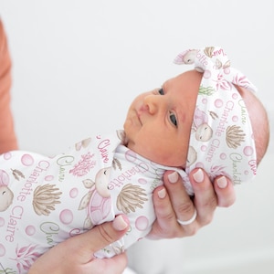 May include: A white swaddle with a pink and green sea turtle pattern and the name "Charlotte Claire" repeated throughout. A baby is wearing the swaddle and a matching headband.