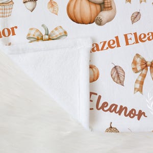 May include: White baby blanket with a fall theme. Features pumpkins, leaves, acorns, cupcakes, and a teddy bear. The name "Eleanor" is printed in orange. The blanket is folded, revealing a soft, white interior.