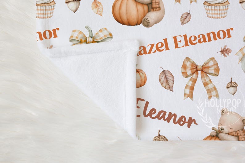 May include: White baby blanket with a fall theme. Features pumpkins, leaves, acorns, cupcakes, and a teddy bear. The name "Eleanor" is printed in orange. The blanket is folded, revealing a soft, white interior.
