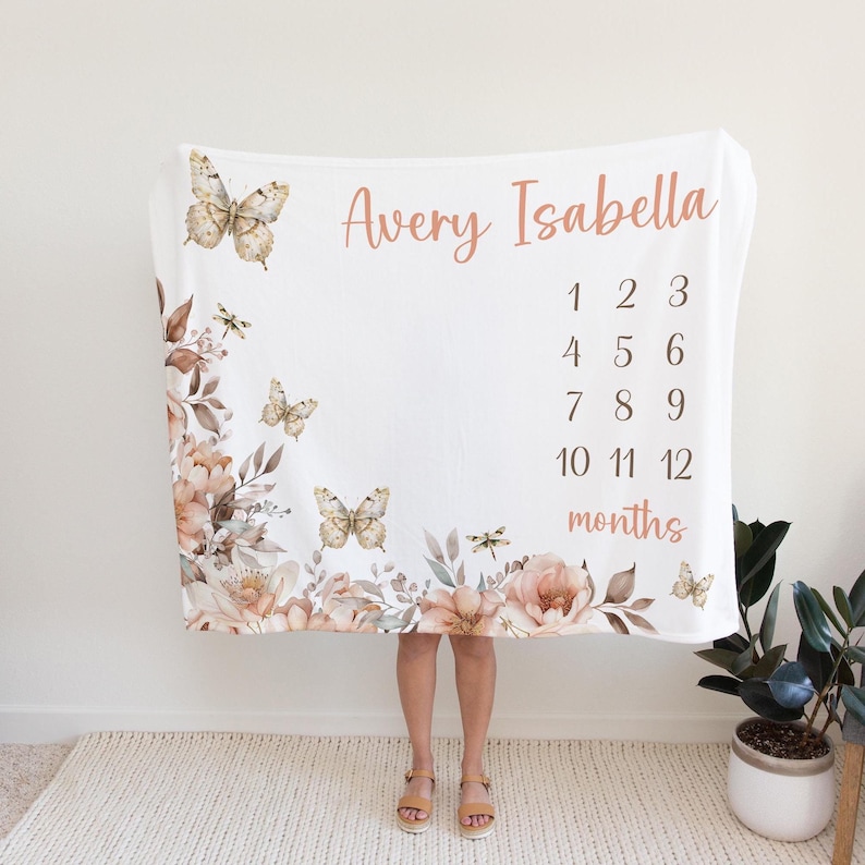 Puede incluir: Una manta blanca para hitos de beb&eacute; con un dise&ntilde;o floral con mariposas y lib&eacute;lulas. La manta tiene los nombres "Avery Isabella" y los n&uacute;meros del 1 al 12 para cada mes del a&ntilde;o.