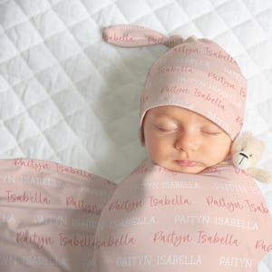 May include: A newborn baby wrapped in a pink swaddle blanket and wearing a matching hat. The blanket and hat are printed with the name "Paityn Isabella" in a repeating pattern. A small teddy bear is next to the baby's face.