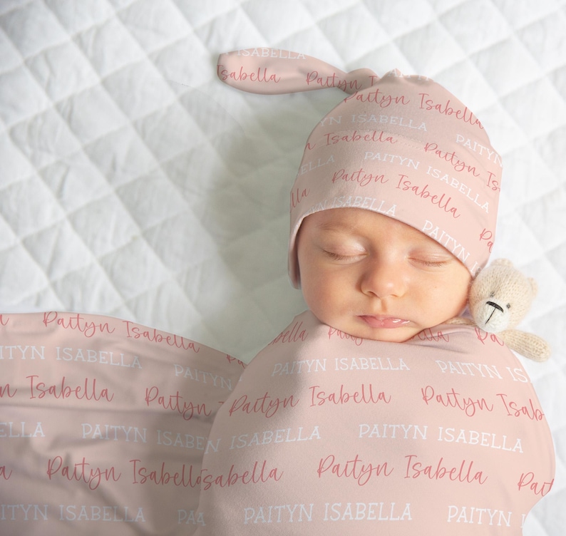 May include: A newborn baby wrapped in a pink swaddle blanket and wearing a matching hat. The blanket and hat are printed with the name "Paityn Isabella" in a repeating pattern. A small teddy bear is next to the baby's face.