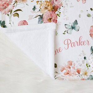 Pode incluir: Um cobertor de beb&ecirc; branco com um design floral com rosas rosa e p&ecirc;ssego, borboletas e o nome "Jane Parker" em escrita rosa. O cobertor tem uma textura macia e felpuda.