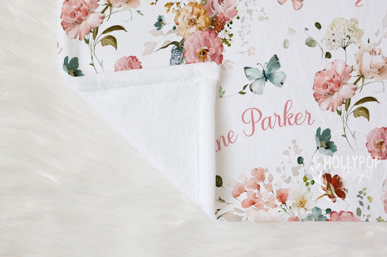 Pode incluir: Um cobertor de beb&ecirc; branco com um design floral com rosas rosa e p&ecirc;ssego, borboletas e o nome "Jane Parker" em escrita rosa. O cobertor tem uma textura macia e felpuda.