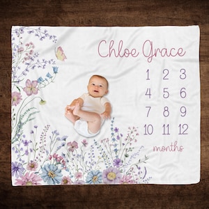 May include: A white baby milestone blanket with floral and butterfly illustrations in shades of purple, pink, and blue. The blanket features the name "Chloe Grace" and numbers 1-12 with the word "months". A baby is lying on the blanket.