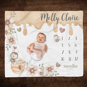 May include: A white baby blanket with a honey drip design and a beehive with a bee. The blanket also has flowers and hearts. The text "Molly Claire" is at the top of the blanket. Numbers 1-12 are printed below the beehive, with the word "months" below the numbers.