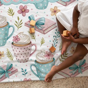 May include: A white blanket with a whimsical pattern featuring coffee cups, books, and floral elements. The cups are in pastel colors with whipped cream and flower accents. A child is playing with wooden blocks on the blanket.