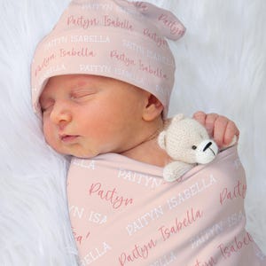 May include: A newborn baby wrapped in a pink blanket and wearing a matching hat, both printed with the name "Paityn Isabella". A small, cream-colored teddy bear is tucked in with the baby. The background is a soft, white, fluffy texture.