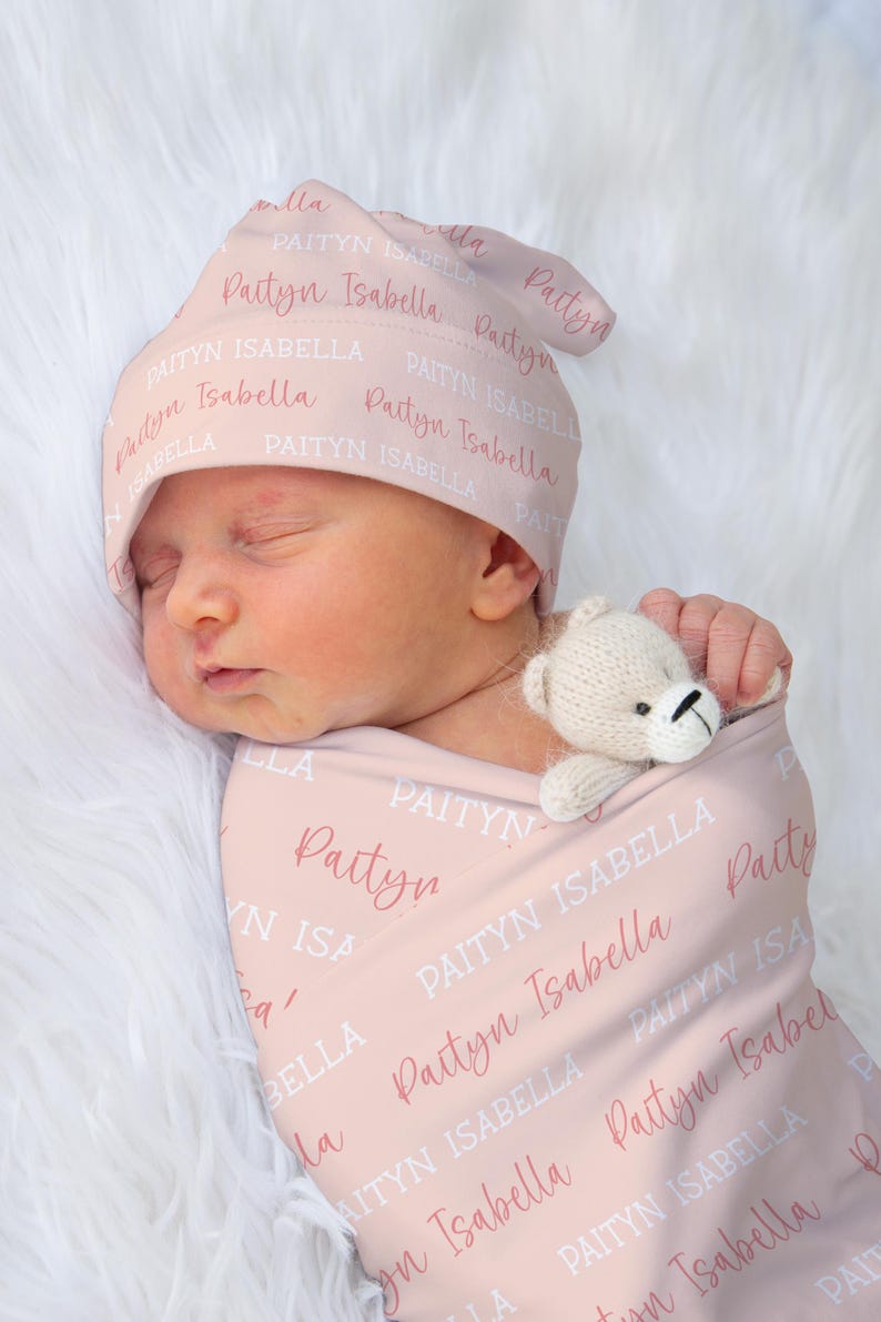 May include: A newborn baby wrapped in a pink blanket and wearing a matching hat, both printed with the name "Paityn Isabella". A small, cream-colored teddy bear is tucked in with the baby. The background is a soft, white, fluffy texture.