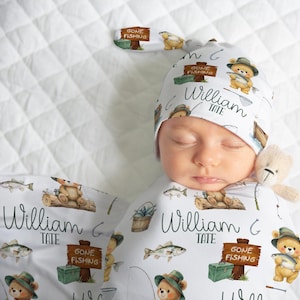 May include: A baby wearing a white swaddle and matching hat, both adorned with a fishing-themed pattern. The design includes teddy bears, fish, fishing gear, and the text "Gone Fishing" and "William Tate". A small teddy bear is nestled near the baby's face.