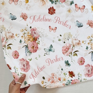 Pode incluir: Cobertor branco com um design floral com flores rosa, p&ecirc;ssego e bord&ocirc;, juntamente com borboletas em tons de azul e rosa. O nome "Adeline Parker" est&aacute; escrito em rosa no cobertor.