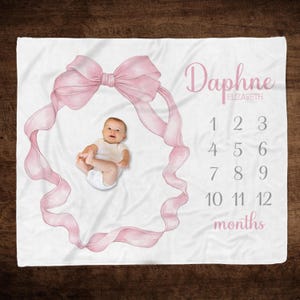 May include: White baby milestone blanket with a pink ribbon bow design. The blanket features the name "Daphne" and numbers 1-12 for monthly photos. A baby is lying inside the ribbon frame.