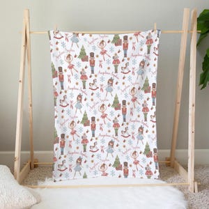 May include: A white blanket featuring a repeating pattern of nutcrackers, ballerinas, Christmas trees, rocking horses, and snowflakes. The name Angelina Sophia is printed in a cursive script throughout the design in a soft pink color.