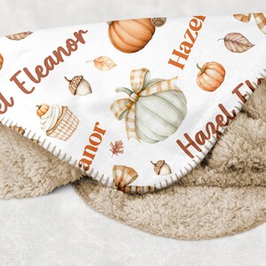 May include: A cozy, personalized baby blanket featuring a white top layer adorned with watercolor pumpkins, leaves, and acorns. The names "Eleanor" and "Hazel" are printed in a warm brown font. The blanket has a soft, tan sherpa backing.