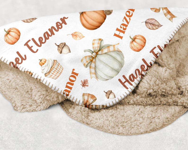 May include: A cozy, personalized baby blanket featuring a white top layer adorned with watercolor pumpkins, leaves, and acorns. The names "Eleanor" and "Hazel" are printed in a warm brown font. The blanket has a soft, tan sherpa backing.