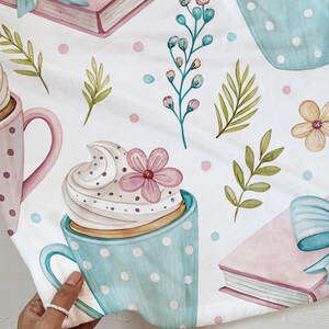 May include: A white fabric with a watercolor pattern featuring light blue polka-dotted mugs with whipped cream and pink flowers, pink books with blue bows, and green leaves. The design also includes pink and blue polka dots.