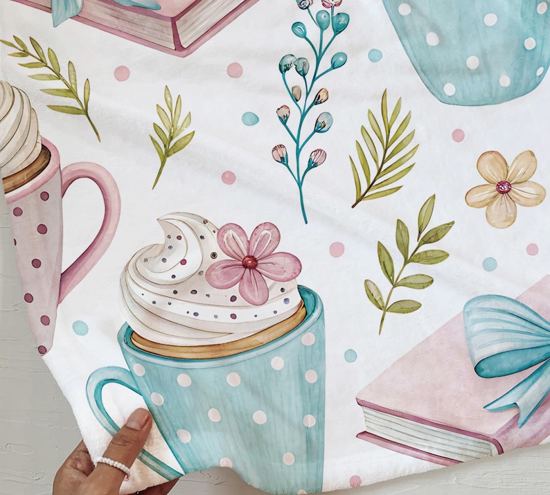 May include: A white fabric with a watercolor pattern featuring light blue polka-dotted mugs with whipped cream and pink flowers, pink books with blue bows, and green leaves. The design also includes pink and blue polka dots.
