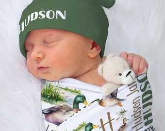 May include: A white swaddle blanket with a green, brown, and white duck pattern. The blanket has the name "Hudson James" written on it. A baby is wearing a green beanie with the name "Hudson" written on it. The baby is holding a small, white teddy bear.