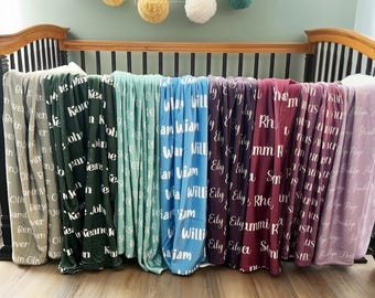 May include: A collection of personalized blankets in various colors, each displaying a name in white script. The blankets are draped over a crib, showcasing a range of hues from gray to purple.
