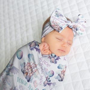 May include: A sleeping baby wrapped in a soft, patterned blanket and wearing a matching bow headband. The blanket and bow feature a whimsical dragon and castle design in pastel colors. The name "Rowan Jade" is printed on the blanket.