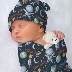 May include: A baby swaddled in a blue and white swaddle with a rocket, astronaut, and stars pattern. The swaddle has the name "William James" repeated throughout the pattern. The baby is wearing a matching blue and white hat with the same pattern.