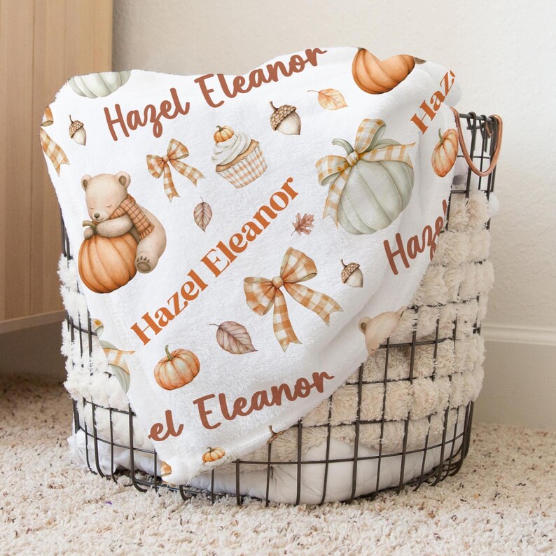 May include: White baby blanket with a fall theme, featuring pumpkins, acorns, leaves, and a teddy bear. The blanket has the name "Hazel Eleanor" printed in orange. The blanket is folded and placed in a black wire basket.
