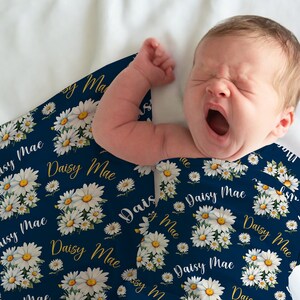 K&ouml;nnte beinhalten: Ein Baby, das in eine marineblaue Musselin-Wickeldecke mit einem wei&szlig;en G&auml;nsebl&uuml;mchen- und goldenen "Daisy Mae"-Druck gewickelt ist.