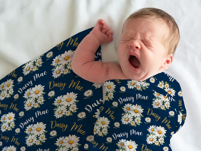 K&ouml;nnte beinhalten: Ein Baby, das in eine marineblaue Musselin-Wickeldecke mit einem wei&szlig;en G&auml;nsebl&uuml;mchen- und goldenen "Daisy Mae"-Druck gewickelt ist.