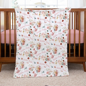 Pode incluir: Um cobertor de beb&ecirc; branco com um padr&atilde;o floral de rosas rosa e p&ecirc;ssego, folhas verdes e borboletas azuis. O nome "Kdelina Parker" &eacute; repetido no cobertor em uma fonte rosa. O cobertor est&aacute; sobre um ber&ccedil;o de madeira.