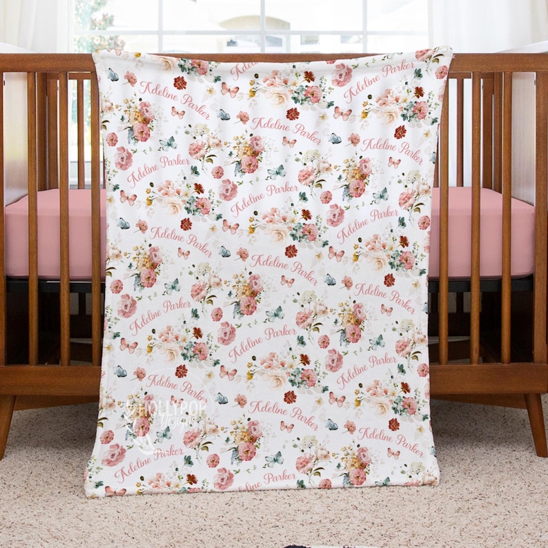 Pode incluir: Um cobertor de beb&ecirc; branco com um padr&atilde;o floral de rosas rosa e p&ecirc;ssego, folhas verdes e borboletas azuis. O nome "Kdelina Parker" &eacute; repetido no cobertor em uma fonte rosa. O cobertor est&aacute; sobre um ber&ccedil;o de madeira.