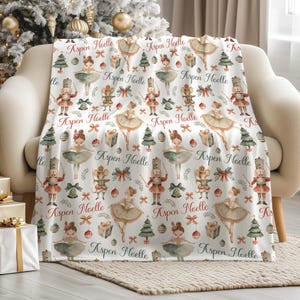 May include: A white fleece blanket featuring a repeating Christmas pattern of nutcrackers, ballerinas, mice, pine trees, and gift boxes. The text Aspen Noelle is printed in a red script font throughout the design, interspersed with small bows.