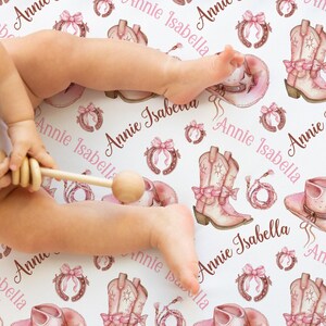 May include: A white baby crib sheet with a repeating pattern of pink cowboy boots, hats, horseshoes, and bows. The name "Annie Isabella" is written across the sheet in a mix of pink and brown fonts. A baby's legs and a wooden rattle are visible.