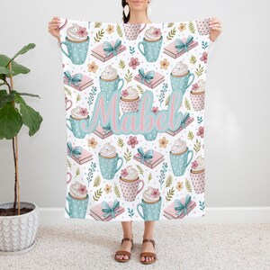 May include: A white blanket featuring a repeating pattern of blue polka-dotted mugs with whipped cream, pink books with blue bows, and floral accents. The name "Mabel" is written in pink and blue script across the center.