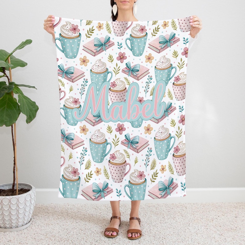 May include: A white blanket featuring a repeating pattern of blue polka-dotted mugs with whipped cream, pink books with blue bows, and floral accents. The name "Mabel" is written in pink and blue script across the center.