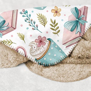 May include: A cozy, white blanket with a watercolor design featuring pink books tied with blue bows, a blue polka-dot teacup with a cupcake, and floral accents. The blanket has a soft, tan sherpa backing.