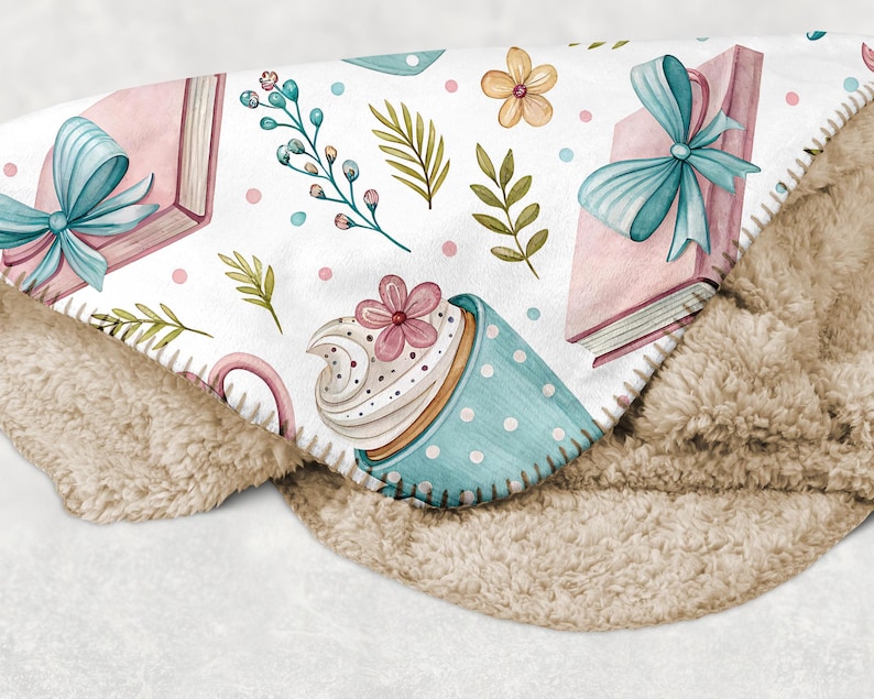 May include: A cozy, white blanket with a watercolor design featuring pink books tied with blue bows, a blue polka-dot teacup with a cupcake, and floral accents. The blanket has a soft, tan sherpa backing.