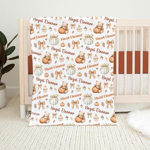 May include: White baby blanket with a repeating pattern of pumpkins, teddy bears, bows, cupcakes, and leaves. The name "Hazel Eleanor" is printed throughout the design. The blanket is draped over a wooden crib.