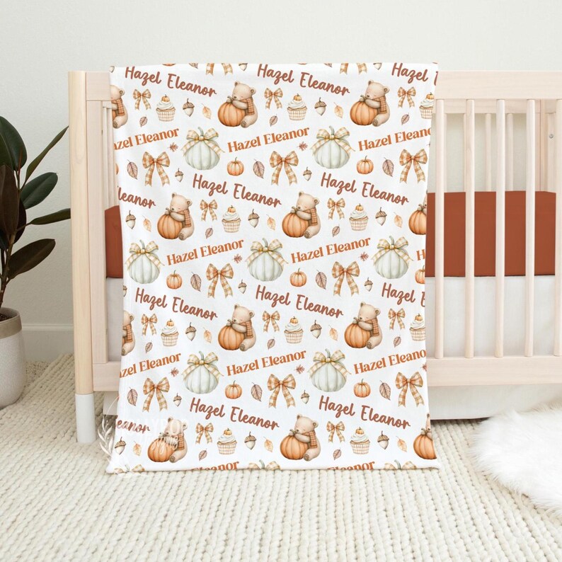May include: White baby blanket with a repeating pattern of pumpkins, teddy bears, bows, cupcakes, and leaves. The name "Hazel Eleanor" is printed throughout the design. The blanket is draped over a wooden crib.