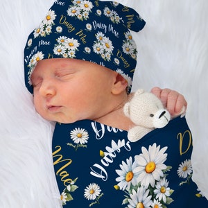 K&ouml;nnte beinhalten: Ein marineblauer Babyschlafsack mit einem wei&szlig;en G&auml;nsebl&uuml;mchenmuster und dem Namen "Daisy Mae" in goldenen Lettern. Der Schlafsack ist um ein schlafendes Baby gewickelt, das eine passende marineblaue und wei&szlig;e G&auml;nsebl&uuml;mchenm&uuml;tze tr&auml;gt.