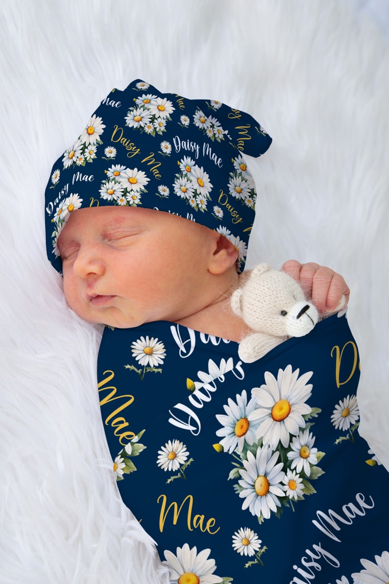 K&ouml;nnte beinhalten: Ein marineblauer Babyschlafsack mit einem wei&szlig;en G&auml;nsebl&uuml;mchenmuster und dem Namen "Daisy Mae" in goldenen Lettern. Der Schlafsack ist um ein schlafendes Baby gewickelt, das eine passende marineblaue und wei&szlig;e G&auml;nsebl&uuml;mchenm&uuml;tze tr&auml;gt.
