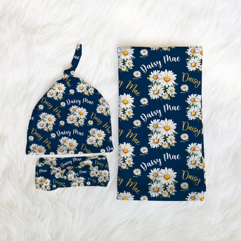 K&ouml;nnte beinhalten: Ein Set aus Baby-M&uuml;tze, Stirnband und Wickeltuch in Marineblau mit einem wei&szlig;en G&auml;nsebl&uuml;mchen- und goldenen "Daisy Mae"-Druck.