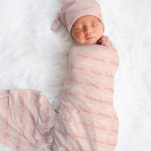 May include: A newborn baby wrapped in a light pink swaddle and matching cap. The fabric is covered in the name "Paityn Isabella" in a repeating pattern. The baby is lying on a white, fluffy surface.