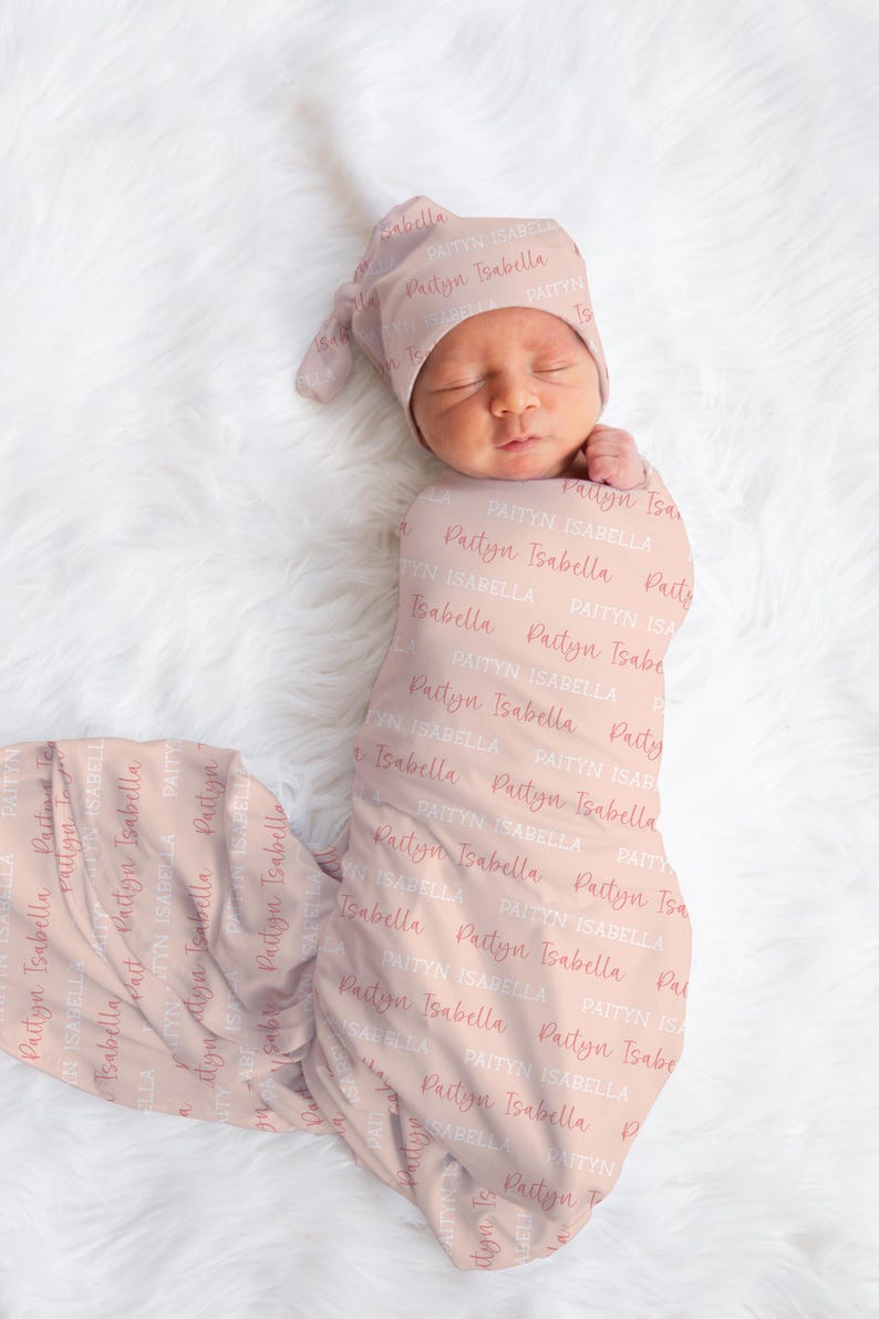 May include: A newborn baby wrapped in a light pink swaddle and matching cap. The fabric is covered in the name "Paityn Isabella" in a repeating pattern. The baby is lying on a white, fluffy surface.