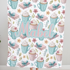 May include: A white blanket with a repeating pattern of blue polka-dot mugs, pink cupcakes, pink books with blue bows, and flowers. The name "Mabel" is written in pink and blue script across the center.