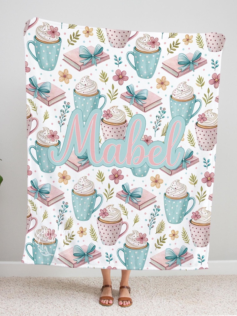 May include: A white blanket with a repeating pattern of blue polka-dot mugs, pink cupcakes, pink books with blue bows, and flowers. The name "Mabel" is written in pink and blue script across the center.