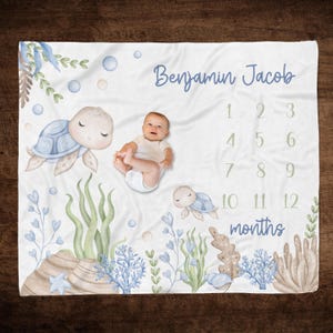 May include: A white baby blanket with a blue and green underwater theme. It features two sea turtles, seaweed, and bubbles. The blanket has a personalized name "Benjamin Jacob" and a number chart for tracking a baby's growth from 1 to 12 months.