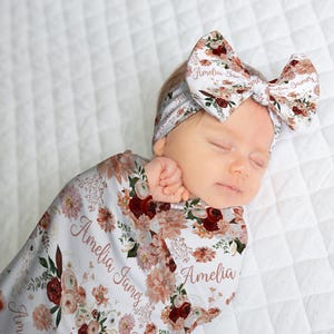 May include: A newborn baby wrapped in a floral swaddle and matching headband. The swaddle and headband feature a white background with burgundy, peach, and green floral patterns. The name "Amelia James" is printed on the swaddle.