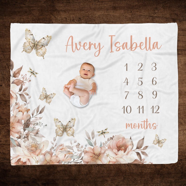 Puede incluir: Una manta de beb&eacute; blanca con un dise&ntilde;o floral y mariposas. La manta tiene una combinaci&oacute;n de colores rosa y marr&oacute;n e incluye una tabla de n&uacute;meros para hacer un seguimiento del crecimiento de un beb&eacute;. El texto "Avery Isabella" y "meses" est&aacute; impreso en la manta.