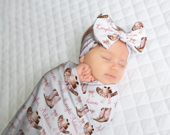May include: A newborn baby wrapped in a light gray swaddle blanket and matching headband. The blanket and headband feature a repeating pattern of cowboy boots, hats, and bows in shades of brown and pink. The name "Layla Addison" is printed on the items.