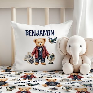 May include: White pillow with the name "BENJAMIN" in navy blue. The pillow features a teddy bear illustration wearing a jacket, vest, and pants, with a globe, airplane, and ABC blocks. A white stuffed elephant is on the right.